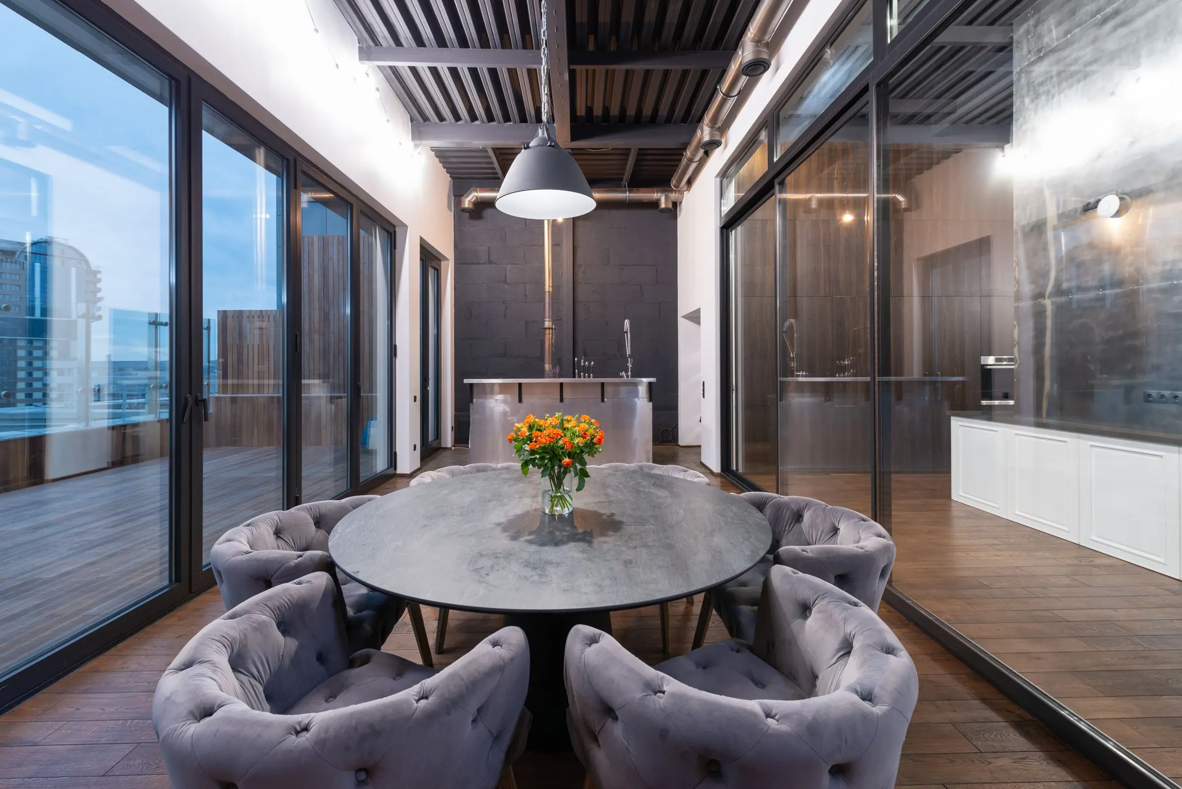 A lounge with a round table and armchairs beside floor-to-ceiling windows in the evening